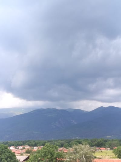 The image depicts a landscape scene with a village in the foreground, nestled against a backdrop of forested hills and mountains under a cloudy sky. The village consists of numerous houses with red-tiled roofs, interspersed with green trees. The houses appear to be single-family dwellings, with some exhibiting small yards and fences. The middle ground is dominated by a dense line of green trees, forming a transition zone between the village and the mountains. The mountains in the background are covered in lush vegetation, with some rocky outcrops visible on their slopes. The sky is filled with heavy, dark clouds, suggesting overcast or stormy weather. A faint object, possibly a bird or a piece of debris, is visible in the sky in the upper left quadrant. The overall impression is one of a rural setting in a mountainous region. The scene is characteristic of Gorno Sahrane, Bulgaria, a village situated in a valley with surrounding hills and mountains.