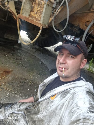 A man wearing a black baseball cap with a headlamp attached, a black t-shirt, and a dirty white hooded jacket is taking a selfie. He has a cigarette in his mouth and smudges of dirt on his face. He is sitting or lying down next to a large, heavy-duty piece of machinery, possibly construction equipment, which is weathered and rusty. The ground is rough and appears to be dirt and gravel, with some green vegetation visible in the background. The overall environment suggests an industrial or outdoor work site, possibly in Zheleznogorsk-Ilimsky, Russia, given the context provided. The image is framed from a low angle, emphasizing the machinery and the man's close-up portrait.
