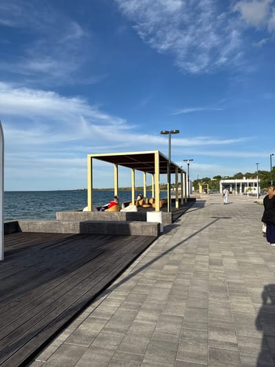 The image captures a modern waterfront promenade in Taman, Russia, bathed in bright daylight under a clear blue sky with scattered wispy clouds. The scene is viewed from a slightly elevated perspective, looking along the length of the promenade towards the horizon.

In the foreground, a paved walkway of grey rectangular stones extends from the bottom right towards the distance. To the left of this walkway, a wooden deck runs parallel to the water. A distinctive yellow-framed canopy or shelter stands over a section of the wooden deck, offering shade. Beneath this shelter, several people are seated, appearing to relax and enjoy the view of the vast body of water. One person is visible wearing a bright red top.

Further down the promenade, towards the middle ground, another person is seen walking away from the camera. Tall, modern streetlights with dual circular fixtures are spaced along the promenade. In the far distance, on what appears to be a green hillside, several white buildings are visible, suggesting a settlement or resort area.

The sun appears to be low in the sky, possibly late afternoon or early morning, casting long shadows across the grey paving stones, notably the shadow of the person in the immediate foreground and the shadows of the yellow structure's supports. The water is calm and blue, reflecting the sky. No discernible text is visible in the image. The overall atmosphere is tranquil and inviting, typical of a coastal leisure area.