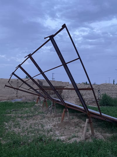 The image captures a rural outdoor scene in Qila Saifullah, Pakistan, dominated by a large, rusty metal framework. This structure, likely designed to hold solar panels, leans across the frame, with some solar panels partially visible at its lower right edge. It is supported by several concrete or wooden posts. The ground beneath and around the structure is a mix of bare, dry soil and patches of low-growing green vegetation, suggesting an agricultural or semi-arid environment.

In the midground, beyond the metal frame, lie remnants of old mud or stone structures, possibly dilapidated buildings or walls, blending into the landscape. Further in the distance, electrical poles and power lines can be seen against the horizon, along with a few sparse trees or bushes.

The sky is heavily overcast with muted blue-gray clouds, indicating either early morning, late afternoon, or a heavily cloudy day. The soft, diffused lighting contributes to a subdued atmosphere. No individuals or discernible activities are present. The scene evokes a sense of neglect or an unfinished project amidst a rustic setting. There is no visible text.