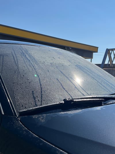 The image shows a close-up of a dark-colored car's windshield, covered in numerous small water droplets and streaks, likely from recent rain or dew. The sun is shining brightly, creating a strong glare on the windshield and highlighting the water. In the background, a yellow and grey structure with a blue metal fence is visible against a clear blue sky. The scene appears to be an outdoor setting, possibly a street or parking area in Velyki Hai, Ukraine. There are no people or visible animals in the frame. The focus is on the car's windshield and the environmental conditions affecting it.