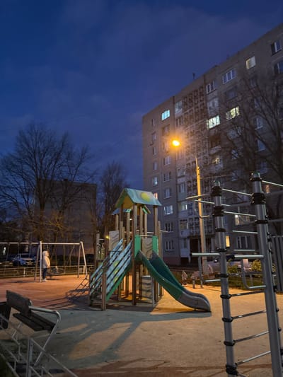 This image depicts a residential playground in Voskresensk, Russia, at twilight or night. A solitary individual, wearing a light-colored hooded coat, stands near a set of metal swings on the left side of the dirt-surfaced playground. In the center, a multi-component playground structure with a light green and yellow roof, wooden supports, stairs, a climbing net, and a dark green slide is present. To the right, a metal climbing frame with grey vertical posts and black horizontal bars is partially visible. A bright orange-yellow streetlamp, positioned in front of a multi-story grey residential building, illuminates the scene, casting long shadows across the playground. The building, prominent on the right, features numerous illuminated windows and a patterned facade. Bare deciduous trees are visible in the left background. The sky is a deep, dark blue with visible cloud formations.