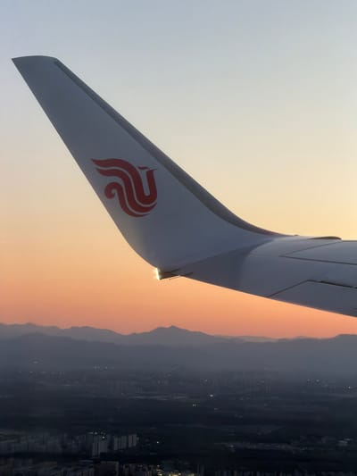 The image depicts the vertical stabilizer of an Air China aircraft in flight. The stabilizer is white with a red stylized phoenix logo. In the background, there's a view of what appears to be a landscape in Shunyi District, China with city lights and mountains visible through a hazy atmosphere. The time of day seems to be dusk or dawn, indicated by the orange-yellow sky.