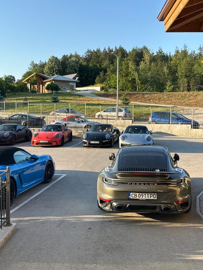In Kerenite, Bulgaria, a parking lot is filled with Porsches. The cars are parked in rows, with some facing forward and others facing backward. In the foreground, a blue Porsche is parked on the left, and a dark green Porsche is parked on the right. In the middle ground, a red Porsche is parked in the center, with black and silver Porsches on either side. In the background, there are several other cars parked, including a white hatchback and a blue hatchback. The parking lot is surrounded by a fence and trees. A modern building with a wooden roof is visible behind the fence. The sky is clear and blue.