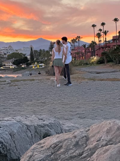 Two individuals, a male and a female, stand on a pebble beach facing away from the camera. The female, with blonde hair pulled back, wears a white tank top and a short green skirt. The male, with dark hair, is dressed in a white t-shirt and dark pants. They appear to be interacting or looking at the scenery.

The background features a vibrant sunset with hues of orange and pink in the sky, casting a warm glow over the scene. In the middle ground, there are buildings with terracotta roofs, palm trees, and some vegetation. A mountain range is visible in the far distance, silhouetted against the colorful sky. The foreground is dominated by a rocky, pebbled shoreline. The overall setting suggests a coastal location, consistent with El Velerín, Spain.