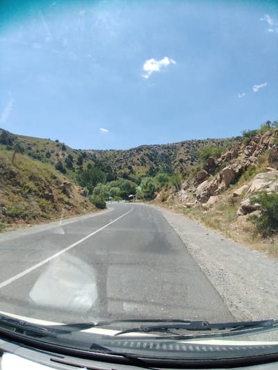 This is a first-person perspective, likely from a vehicle, looking out at a winding road leading through a mountainous, arid landscape. The road is paved with a white line dividing the lanes, and a gravel shoulder runs alongside. The terrain is characterized by dry, scrubby vegetation and rocky outcrops, with sparse green trees dotting the hillsides. In the distance, a small cluster of buildings with green roofs is visible near the bend in the road. The sky is a clear blue with a few scattered white clouds. The sun appears to be high, suggesting it is midday. There are no visible people or specific objects of interest, aside from the natural scenery and the road itself. The overall impression is of a journey through a remote, sun-drenched environment.