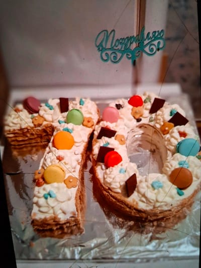 This image captures a decorative cake shaped like the number "70" in the city of Medvezhyi Ozyora, Russia. The cake is covered in white frosting and adorned with colorful macarons, chocolate pieces, and small flower-shaped candies. A turquoise decorative topper with Cyrillic lettering, likely "Поздравляю" (Congratulations), is placed on the cake. The cake sits on a silver foil-covered surface. The background is blurred, suggesting an indoor setting. There are no people visible in the image. The focus is entirely on the cake and its decorations, indicating a celebratory occasion. The presence of the cake suggests a party or gathering in Medvezhyi Ozyora.