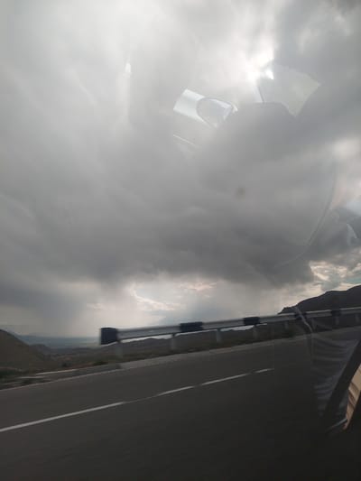 The image captures a dramatic outdoor scene, likely taken from inside or very close to a moving vehicle. The dominant feature is a heavily clouded sky, with thick grey clouds interspersed with areas where brilliant sunlight breaks through, creating strong highlights and lens flares, particularly on the left side.

In the upper-left portion, reflections are visible on what appears to be a car window or sunroof. These reflections include distorted shapes of the clouds and possibly elements of the car's interior, such as a sun visor or dashboard.

To the right, a paved road with a clear white line extends into the distance. Alongside the road, a rugged, mountainous, or hilly landscape is visible, suggesting a dry or arid environment. A metallic structure, possibly a roof rack or part of the vehicle's exterior, is present along the right edge of the frame.

The time of day appears to be daytime, characterized by intense sunlight despite the heavy cloud cover. The weather is dynamic, with bright, breaking sun through an otherwise overcast sky. The location context of Tigranashen, Armenia, aligns with the visible mountainous terrain. No people or visible text are present in the image. The scene evokes a sense of travel and observation of the vast, natural landscape under striking weather conditions.