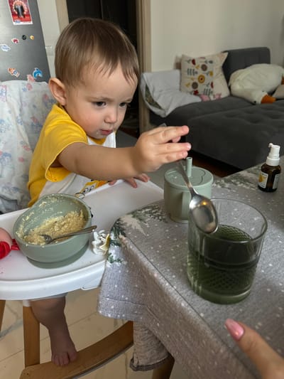 The image depicts an indoor scene in Nikolina Gora, Russia. A young child, identified as a toddler with light brown hair, is seated in a white high chair with a patterned fabric lining. The child wears a yellow t-shirt over a white t-shirt, and food residue is visible on their face. The child's right arm is extended, with the hand reaching towards an adjacent table.

On the high chair's tray, a light green bowl contains a light-colored, semi-solid food, with a spoon resting in it. A white hair claw clip is also present on the tray. The table is covered with a grey tablecloth featuring a white winter-themed pattern. On the tablecloth, a light green sippy cup with a lid and a straw hole is positioned. A metal spoon is horizontally balanced across the rim of the sippy cup and a clear glass tumbler. The glass contains a dark green liquid. To the right of the sippy cup, a small amber spray bottle with a white pump dispenser is present. In the bottom right corner of the frame, an adult's hand with pink manicured fingernails is visible, pointing towards the glass.

The background shows a dark refrigerator adorned with various magnets, including one depicting a person, to the left. In the mid-background, a dark grey sofa is visible, topped with several decorative cushions, one featuring a floral pattern, and a large white goose-shaped plush toy. The room has light-colored walls and flooring.