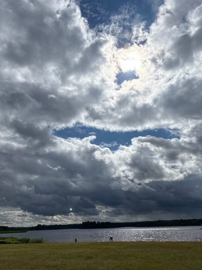 The image depicts a partly cloudy sky over a body of water, likely a lake or river, with a grassy bank in the foreground. The sun is visible through a break in the clouds, casting bright rays. On the grassy bank, there are a few scattered figures, one of whom is wearing a red garment. The water's surface is sparkling, suggesting sunlight reflecting off the ripples. The horizon is lined with trees. The scene appears to be outdoors in a natural setting, possibly a park or recreational area, given the presence of the water and the open grassy space. The overall impression is of a dynamic sky with contrasting light and shadow.