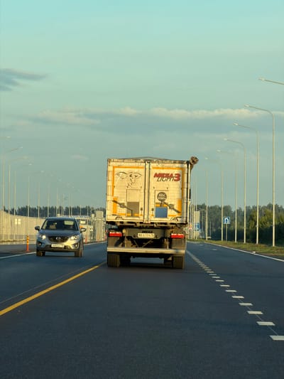 In Kaipy, Russia, a truck and a car are traveling on a highway. The truck, a large white vehicle with "MEGA LIGHT 3" written on its side, is in the right lane. A car, a silver SUV, is in the left lane behind the truck. The highway is flanked by streetlights and trees. The sky is blue with some clouds. The road has yellow and white lines.