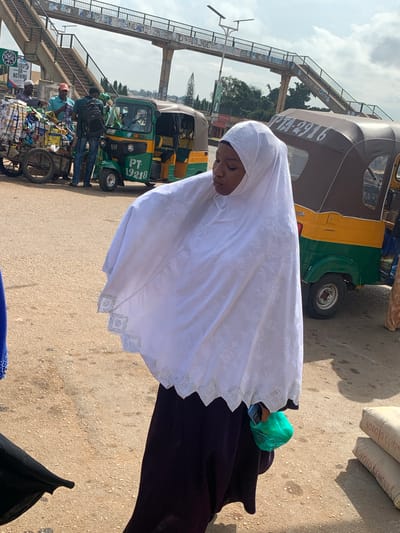 In Gura Top, Nigeria, a young woman in a white hijab and dark purple dress stands on a dirt road. She holds a green plastic bag and her head is bowed. Behind her, a yellow and green auto-rickshaw is parked, displaying the license plate PTA-2916. Further in the background, a pedestrian overpass spans the road, with signs visible underneath. To the left, a cart filled with various goods is being attended to by several men. Bags of "Sokoto Cement" are stacked on the right side of the image. The scene appears to be a busy marketplace area with elements of urban infrastructure.