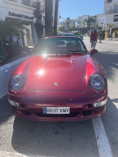 A metallic red Porsche 911 is parked on a sunlit street in Nueva Andalucía, Spain. The car's front end, including its distinctive headlights and emblem, is the primary focus. The license plate, bearing the numbers "8637 KMV", is clearly visible. In the background, a building with signage for "Sabadell Solbank" is partially obscured by greenery. Pedestrians can be seen walking along the sidewalk and crossing the street, suggesting a lively urban environment. The scene is characterized by bright sunlight, casting shadows and creating reflections on the car's polished surface.