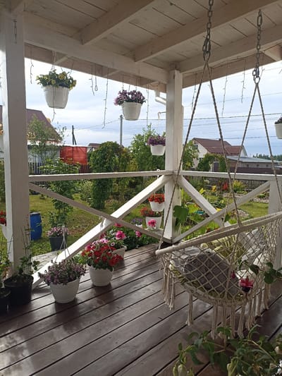 A bright, sunlit image shows a wooden porch with a hanging hammock chair. The porch, constructed of light-colored wood, features a white railing adorned with various potted flowers. Among the blooms are red, pink, and purple petunias, as well as yellow flowers. The hammock chair, made of macrame, is suspended by chains from the porch ceiling and contains cushions. Beyond the porch, a lush green garden is visible with trees, shrubs, and a fence. In the background, residential buildings and a red storage shed are present. The sky above is overcast with clouds. The scene appears to be a tranquil domestic setting, likely a private residence in a suburban or rural area.