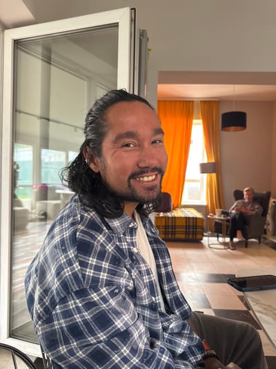 An indoor shot depicts a man with long, dark hair and a beard smiling at the camera, wearing a blue and white plaid shirt. He is positioned in the foreground, with a portion of a glass door visible to his left. In the background, a woman sits in an armchair, looking at a phone. The room features orange curtains, a patterned ottoman, and two pendant lights. The floor is tiled with a checkerboard pattern. This scene appears to be a residential interior. The location provided is Ptichnoye, Russia.