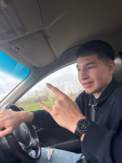 A young man sits in the driver's seat of a car, presumably a Honda, as indicated by the steering wheel logo. He wears a black, zippered jacket and jeans with a distressed or ripped knee. He has a digital watch on his left wrist showing "11:37". The man is pointing to the windshield with his right hand. The environment visible through the windshield appears to be a rural area with trees, likely taken during overcast daylight. There is no evidence in the provided image to ascertain whether the image was taken in Spahievo, Bulgaria.
