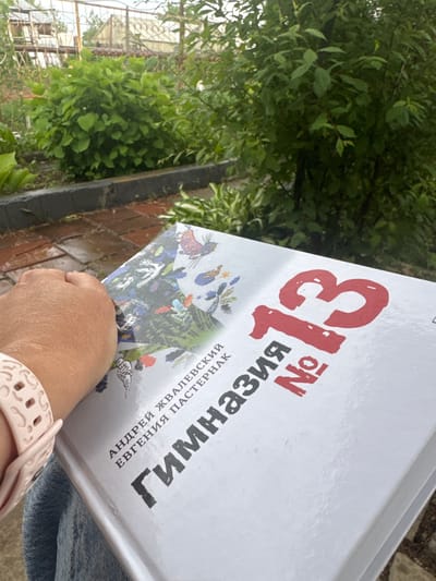 In Ptichnik, Russia, a person holds a white book with a large red number "13" on its cover. The book also features text in Cyrillic script, including "Гимназия No" and authors' names "Андрей Жвалебский" and "Евгения Пастернак". The cover is illustrated with colorful drawings of animals and plants. The person is wearing a light pink watch on their left wrist and denim clothing. The scene appears to be outdoors, with lush green foliage and trees in the background, suggesting a garden or park setting. The ground in the foreground is paved with bricks, some of which appear wet. In the far background, structures resembling a greenhouse and fencing are visible. The lighting suggests it is daytime, possibly overcast.