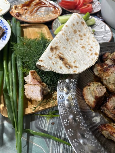 The image shows a close-up of a table laden with food, likely after a meal. In the foreground, a metal platter holds several pieces of cooked meat, appearing to be skewers or grilled chunks, glistening with juices. To the left, a bunch of green onions lies on a wooden cutting board, next to a pile of fresh dill. A piece of flatbread is folded and placed on top of the dill. In the background, partially visible, are plates with remnants of food, including a dish with a reddish sauce and sliced tomatoes and cucumbers. The overall scene suggests a home-cooked meal, possibly a barbecue or a gathering, in a domestic setting. There are no people visible in the image, and no specific details of the location in Подмой, Russia, can be discerned from the visual information provided.