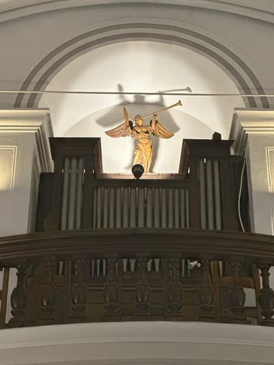 The image shows a carved wooden angel with outstretched wings, holding a trumpet. The angel is positioned above a pipe organ, which is made of dark wood and has many vertical pipes. The organ is behind a wooden railing with decorative balusters. The background is a white, arched wall with shadow lines creating a sense of depth. The lighting appears to be artificial, casting shadows from the angel and the organ pipes. This scene is located within the Durbuy Castle in Durbuy, Belgium.