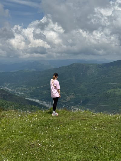 The image shows a person standing on a grassy hill overlooking a valley with mountains. The person is wearing a pink t-shirt, black leggings, a black cap, and white and red sneakers. They are facing away from the camera, looking out at the landscape. The hill is covered in green grass and scattered white wildflowers. In the background, there are several layers of green mountains with a river running through the valley. The sky is cloudy with patches of blue. The scene appears to be a scenic overlook in a mountainous region, possibly in Nizhnyaya Teberda, Russia, given the context provided. A thin wire fence runs horizontally across the middle ground of the image, separating the foreground from the valley.