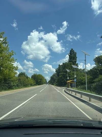 This is a photograph taken from inside a vehicle, looking out at a road and its surroundings. The scene is dominated by a bright blue sky with scattered white, fluffy clouds, suggesting a pleasant, sunny day. The time of day appears to be late morning or early afternoon due to the brightness and quality of the light.

The road itself is asphalt, with a white line marking its edge and a dashed white line indicating lanes. On the left side of the road, a metal guardrail runs along the shoulder, separating it from a grassy area and then a dense line of green trees. The trees form a thick, verdant backdrop, with their leaves appearing full and lush.

Strategically placed along the roadside are several poles equipped with solar panels, angled towards the sun. These appear to be part of some form of roadside infrastructure, possibly for lighting or communication. One of these poles also has a yellow structure with a blue sign depicting a bus. This suggests a bus stop is located nearby.

There are no people directly visible in the image, but there are several vehicles further down the road, indicating traffic. The overall impression is one of a rural or semi-rural highway setting, possibly in a temperate climate given the foliage. The location context of Goryachy Klyuch, Russia, further supports this interpretation. The image is oriented vertically, with the road and trees running from the bottom right to the top left of the frame, and the sky occupying the upper left portion.