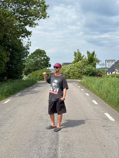 A man is captured in a daylight outdoor shot, standing upright on an asphalt road. He is positioned slightly left of the center, looking towards the camera. He wears a black t-shirt featuring a black and white graphic print of a person's face, with the text "regine lanch" visible below it. He is also dressed in black shorts, black slide sandals, and a maroon baseball cap, with sunglasses resting on his face. His expression appears neutral. He holds a long, thin stick vertically in his right hand, while his left hand rests in his pocket.
The asphalt road has white dashed lines visible on its right edge. A long shadow of the man is cast to his left, indicating the sun is at a moderate angle, possibly in the late afternoon. Lush green grass and trees line both sides of the road.
In the mid-ground on the right, a brown wooden sign is visible. The sign clearly displays white text: "VESSIGEBOD" at the top, followed by "LÖRDAG" and "SÖNDAG" (Swedish for Saturday and Sunday), with the numbers "3-17" below, likely indicating open hours (e.g., 3 PM to 5 PM). Behind the sign, white houses with dark roofs can be seen nestled among green hills and trees. The background features a partly cloudy blue sky. The scene depicts a rural or semi-rural setting, identifiable as Vessigebro, Sweden, based on the sign's text.