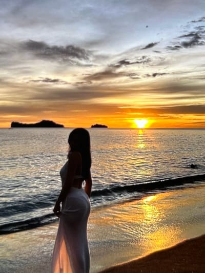 The image depicts a serene sunset scene at a beach. A person, silhouetted and wearing a white dress, stands near the water's edge, facing away from the camera and towards the ocean. The sun is setting on the horizon, casting a warm, golden glow across the water and sky. Islands are visible in the distance. The sand is wet from the receding tide, and small waves are gently lapping at the shore. The sky is a mix of orange, yellow, and grey clouds. The scene is tranquil and evocative of a peaceful evening by the sea. This image is not located in Montfermeil, France.