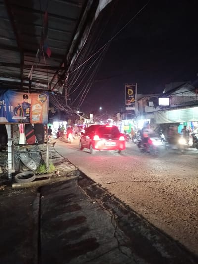 This image captures a nighttime street scene in Ciomas, Indonesia. The vantage point is from a cracked concrete sidewalk on the left, looking down a two-lane asphalt road that curves slightly to the right. The sky overhead is dark.

On the immediate left, a corrugated metal canopy extends over the sidewalk, from which numerous tangled utility wires descend and crisscross the upper portion of the frame. Beneath this canopy, a blue banner featuring an advertisement for "TOP 1 EXTREME" motor oil, depicting a man in a racing suit, is displayed. Below the banner, a discarded tire and what appear to be stacked motorcycle seat parts or similar black objects are visible on the ground.

The road is actively traversed by vehicles. A red passenger car, with its taillights illuminated, is moving away from the viewer in the center of the road, exhibiting motion blur. Numerous motorcycles are also present, both in front of and alongside the red car. Several motorcycles have active headlights and taillights, illuminating parts of the road and other vehicles. Riders are visible on most motorcycles, though they are often blurred due to motion.

On the right side of the road, various commercial buildings line the street, illuminated by their own lighting. A prominent yellow vertical sign with "BAKSO JANGKUNG" and "Rp. 15.000" is visible further down the street on the right. Other indistinct storefronts and blurred human figures are discernible in the background, indicating ongoing pedestrian and commercial activity. The scene suggests a busy urban or semi-urban thoroughfare during evening hours.