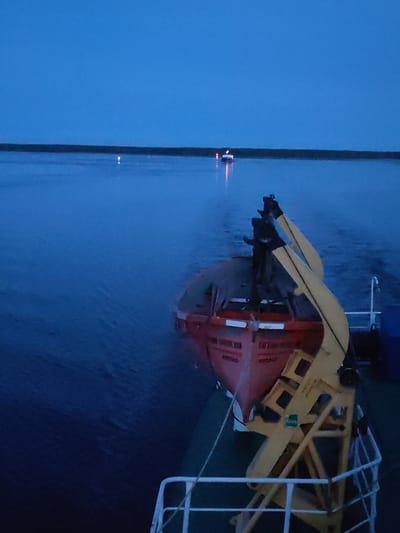 The image shows a view from a ship at dusk. In the foreground, a red lifeboat is visible, suspended by a yellow davit. The boat has some writing on its side, but it is largely unreadable due to the lighting and image quality. The sea is calm and dark blue, with faint ripples. In the distance, a dark horizon line separates the water from the sky. There are a few distant lights visible on the horizon, suggesting a coastline or other vessels. The sky is a deep blue, transitioning to black at the top. The overall impression is one of a serene and somewhat melancholic twilight scene at sea. The location is specified as Malinukha, Russia, which is a small village in the Arkhangelsk Oblast, situated on the coast of the White Sea.