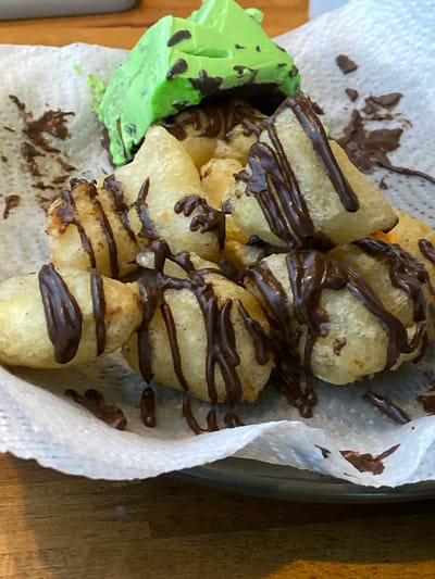 A pile of golden fried pastry pieces, drizzled with dark chocolate sauce, sits on a white paper napkin. A bright green, possibly lime-flavored, dessert or ice cream scoop is placed on top of the pastries. The food is arranged on a dark plate, which is on a wooden table. This scene is located in Candirejo, Indonesia.