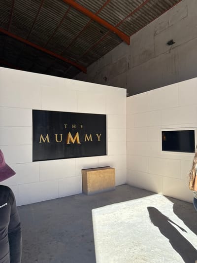 The scene depicts an interior exhibit space, likely within a film studio or museum, located in Ouarzazate, Morocco. The environment features an L-shaped corner formed by white paneled walls, set beneath a high, dark corrugated metal roof supported by reddish-brown trusses. The floor is gray concrete, partially illuminated by strong directional light creating distinct shadows.
Centrally positioned on the main white wall is a black rectangular panel displaying the gold text "THE MUMMY," with a prominent vertical lighter streak on the second 'M'. Below this sign, a light brown, rectangular block rests on the concrete floor. On the adjacent white wall to the right, a smaller black rectangular screen or monitor is recessed.
Two individuals are partially visible: one on the far left, showing a portion of a person wearing a purple baseball cap and a dark long-sleeved shirt with "OLAIAN" text; and another on the far right, indicated by a visible arm, hair, and a long, clear shadow cast across the concrete floor. No direct interactions or specific actions are discernible between the individuals.