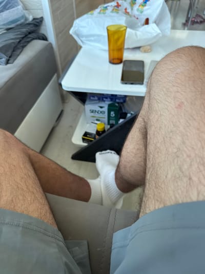 A close-up, first-person perspective shot shows a person sitting on a sofa in what appears to be a living room. The person is wearing grey shorts and white socks. In the background, a small white table is visible, holding a yellow cup, a dark-colored smartphone, and a box containing what looks like medication. A white plastic bag is also on the table. The scene is well-lit, with natural light coming from the right side of the frame. The overall impression is one of a relaxed, domestic setting. The image does not provide any specific clues to indicate its location in Dibich, Bulgaria, or any other city.