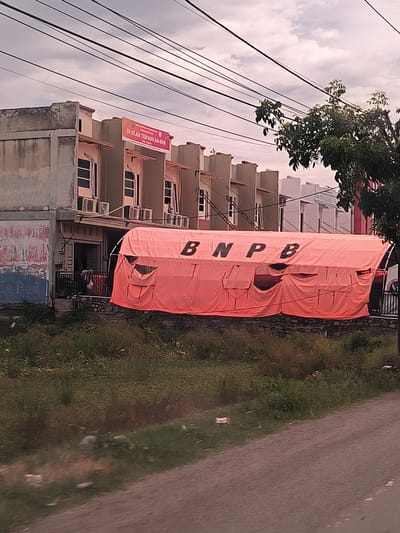 The image captures a street scene in Bunot, Indonesia, featuring buildings, a prominent orange tent, and natural elements under an overcast sky.
In the background, a row of multi-story buildings stands. The building on the far left appears weathered, while others are more uniform and modern, possibly residential or commercial units. A red and orange banner is affixed to one of these buildings, displaying the text "SD ISLAM TERPADU AN-NUR," indicating an integrated Islamic elementary school. Multiple air conditioning units are visible mounted below the windows of these structures.
A large, bright orange tent dominates the midground, positioned in front of the buildings and atop a low stone wall. The tent features the bold black letters "BNPB" prominently on its side, an acronym for Indonesia's National Disaster Management Agency (Badan Nasional Penanggulangan Bencana). Several openings or vents are visible along the tent's lower half. The presence of this tent suggests a temporary facility, potentially for disaster response, relief efforts, or a community event.
The foreground is characterized by an expanse of overgrown green and brown grass and weeds, separating the viewer from the tent and buildings. A dirt or gravel road partially fills the bottom of the frame. Above, numerous dark power lines crisscross the cloudy, light grey sky. A section of a leafy green tree is visible on the right side of the image. The time of day appears to be daytime, though the overcast conditions create soft, diffused lighting. No individuals are visible, and no specific activity is taking place.