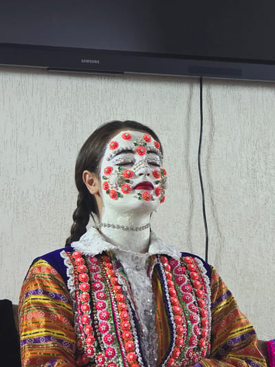 A young woman with her eyes closed is adorned with white face paint and floral and sequined decorations on her face, with her hair in a braid. She wears a traditional, colorful embroidered garment and a beaded necklace. The background consists of a white textured wall and a Samsung television. This image captures a moment from the traditional Kukeri festival in the village of Ribnovo, Bulgaria.