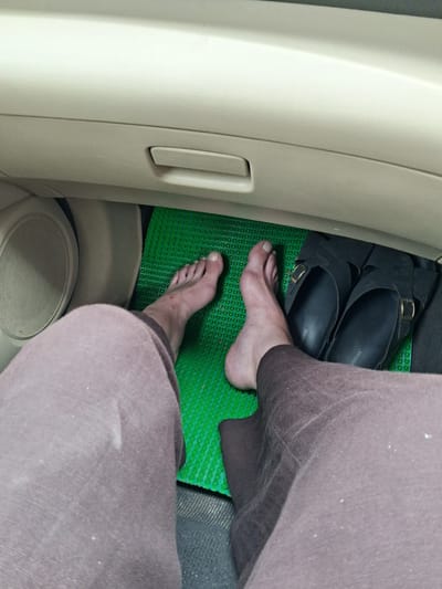 The image shows a person's bare feet and a pair of black sandals placed on a green mat inside a car. The car's interior is visible, including the dashboard and a glove compartment. The scene appears to be located in Nagwain, India. The lighting is somewhat dim, suggesting it might be daytime with overcast skies or the interior of the car is not well-lit. There is no indication of any criminal activity or foul play.