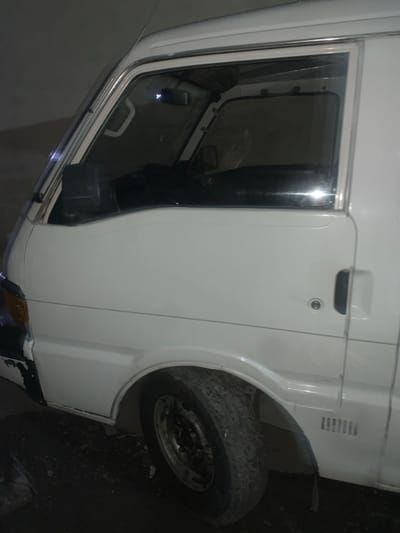 The image shows a white vehicle from a close-up perspective, likely the driver's side door and front wheel. The door features a reflective window, a handle, and a lock. The tire is heavily textured and covered in dirt or grime, suggesting recent use in off-road conditions. The vehicle's body panels appear clean, highlighting a contrast between the pristine exterior and the dirty wheel. Some damage or repair is visible at the bottom of the image.