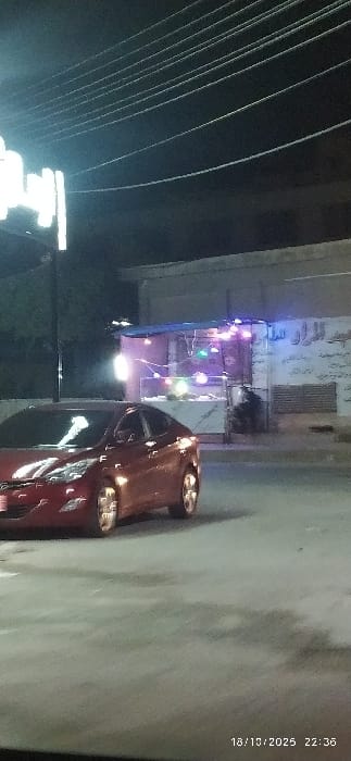 This image, taken at 22:36 on October 18, 2025, depicts a nighttime street scene in Al-Kiswa, Syria. A red sedan, likely a Hyundai Elantra model, is positioned in the lower-left to mid-frame, facing right, with its headlights illuminated. Overhead, numerous utility lines are visible against a dark sky. To the right of the vehicle, a building with a light-colored facade features Arabic script on its wall, suggesting a commercial establishment. In front of this building, an outdoor stall with a blue canopy is set up, illuminated by a string of colorful globe lights. A display case with a glass top is visible at the stall. A solitary individual is partially visible in the shadowed area to the right of the stall, seated or standing. The ground surface appears to be asphalt or concrete. A bright light source or streetlight is present on the far left.