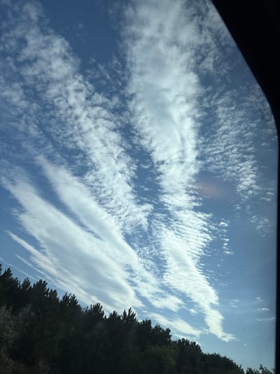 The image shows a view of the sky above a line of trees in Biatorbágy, Hungary. The sky is a bright blue with scattered white clouds. The clouds are wispy and appear to be moving from left to right. The trees are dark green and appear to be pine trees. The image appears to have been taken from a moving vehicle, as indicated by the slight blur in the foreground and the framing of the shot by what looks like a car window. There are no people or animals visible in the image. The overall scene suggests a pleasant day with good weather.