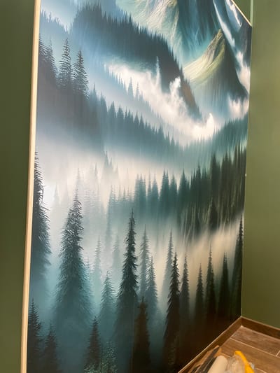 The image displays a large, vibrant mural of a misty forest with majestic mountains in the background. The mural is mounted on a green wall. To the right of the mural, there is a corner where the green wall meets another green wall, and a wooden baseboard runs along the bottom. On the floor in this corner, there are tools likely used for installing the mural, including rolls of material, a scraper, and a yellow object that may be a bag or tarp. The scene is devoid of people. The mural's artistic style is somewhat stylized and evocative, with a focus on atmospheric lighting and deep greens and blues. The overall impression is that of a new installation in progress or recently completed within an interior space. There is no indication or information linking this image to the city of Meaux, France.