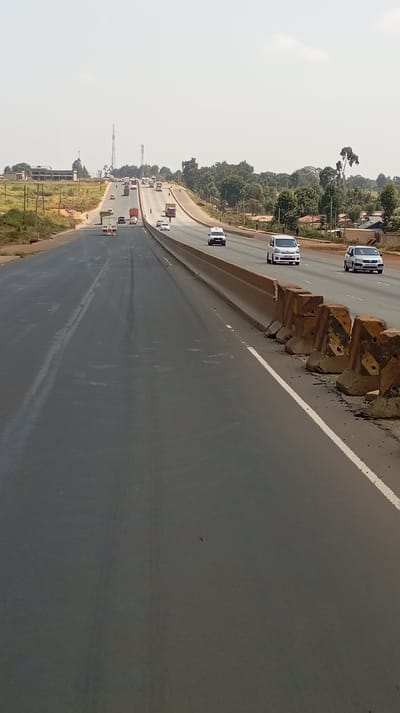 The image displays a multi-lane highway in Muguga ward, Kenya, ascending a gradual incline under a hazy sky. In the immediate foreground, a dark gray asphalt lane, approximately 10 meters wide, appears recently paved, marked by a faded white line on its right edge. This lane is devoid of active traffic. To its right, a continuous light brown concrete median barrier separates it from active traffic lanes. Positioned at the base of this median are several worn, orange-brown concrete Jersey barriers, some appearing displaced or damaged.

Beyond the median, multiple active highway lanes carry vehicular traffic away from the viewer. Vehicles present include several white vans (e.g., Toyota Hiace-type models), a dark sedan, numerous large cargo trucks (including a prominent white semi-truck and several brown flatbed or box trucks), smaller passenger cars, and at least one motorcycle or scooter. No individuals are discernible within or around the vehicles.

To the far left of the highway, an unpaved shoulder or dirt access road runs parallel to the main thoroughfare, lined with utility poles. Beyond this, an elevated, reddish-brown expanse of cleared land or construction ground features a distant multi-story building with a dark roof and light-colored walls, alongside several tall communication towers. The far right side of the highway is bordered by dense green tree cover, partially obscuring scattered residential structures. The sky is uniformly hazy light blue to white, indicating clear weather.