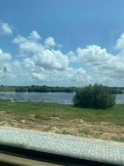 The image shows a view from inside a vehicle, likely a car, looking out at a landscape that includes a body of water, possibly a river or a canal. The sky is visible, with patches of blue and puffy white clouds. Alongside the water, there is green vegetation, possibly grass and trees. A paved or brick-laid surface is visible in the foreground, partially obstructed by the vehicle's frame. The location is identified as the city of Avlékété, Benin. The image does not provide any information about people or specific actions. The water is calm with reflections.