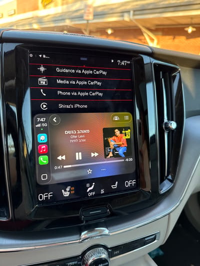 The image displays the interior of a vehicle's dashboard, specifically focusing on the infotainment system and surrounding elements. The scene is located in Lawrence Township, United States.
Central to the image is a black rectangular touchscreen showing an active Apple CarPlay interface. The time displayed is 7:47 AM, with a 5G cellular signal indicator present. The screen lists options such as "Guidance via Apple CarPlay," "Media via Apple CarPlay," and "Phone via Apple CarPlay." Below these, "Shiraz's iPhone" is connected, displaying a music player for the artist "Ofer Levi" and a song titled in Hebrew "מאוהב בגשם." The album art features a man in an orange shirt and blue jeans. The song is 47 seconds into playback with 6 minutes and 20 seconds remaining. Below the music player, climate controls show heated seats are off, fan speed is set to "88" with an "OFF" indicator, and "AC OFF."
To the right of the screen, a vertical air vent with a chrome-trimmed adjustment knob is visible. The dashboard features glossy black plastic trim surrounding the screen and vent, contrasting with a lighter beige/grey lower dashboard panel. A chrome-accented rotary control knob for media playback is partially visible on the center console below the screen.
Through the vehicle's windshield, an out-of-focus exterior environment is visible under bright daylight. The background shows a brick building with a light-colored overhang or canopy structure, from which several recessed light fixtures protrude. A blurred red signpost is also present, suggesting a commercial or retail setting. No individuals are visible within the frame.