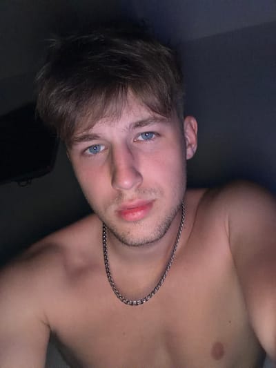 A shirtless young man with short, light brown hair and striking blue eyes looks directly at the camera. He has a light beard and is wearing a silver chain necklace. The background is dark and appears to be indoors, with a hint of a screen or monitor on the left side. The image is a close-up portrait, focusing on the man's face and upper chest. The lighting is dim, creating a moody atmosphere. The image does not provide any information about the city of Vanino, Russia.