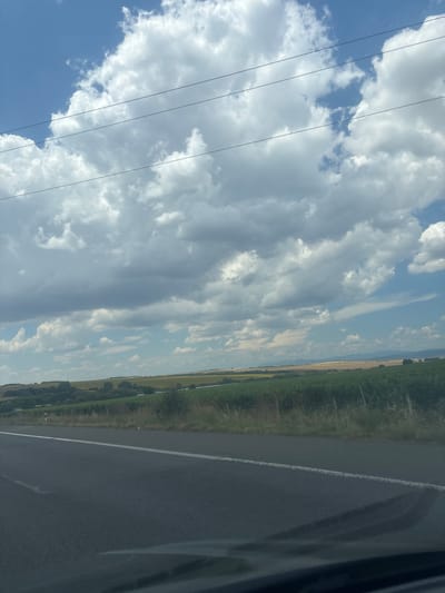 The image shows a scenic view of a rural landscape with a road in the foreground and fields and hills in the background. The sky is filled with large, fluffy white and gray clouds against a bright blue sky. There are power lines running horizontally across the upper portion of the image. The road, which appears to be a paved highway, has a white line marking its edge. To the right of the road, there are fields of green crops, some of which appear to be vineyards or tall bushes, bordered by dry grass and some sparse trees. Beyond the green fields, there are rolling hills with patches of yellow and brown vegetation, suggesting agricultural land. The overall impression is one of a bright, possibly sunny day in a rural area. The location is identified as Zheleznik, Bulgaria. There are no people or any specific actions or interactions depicted in the image. The focus is on the natural environment and infrastructure.