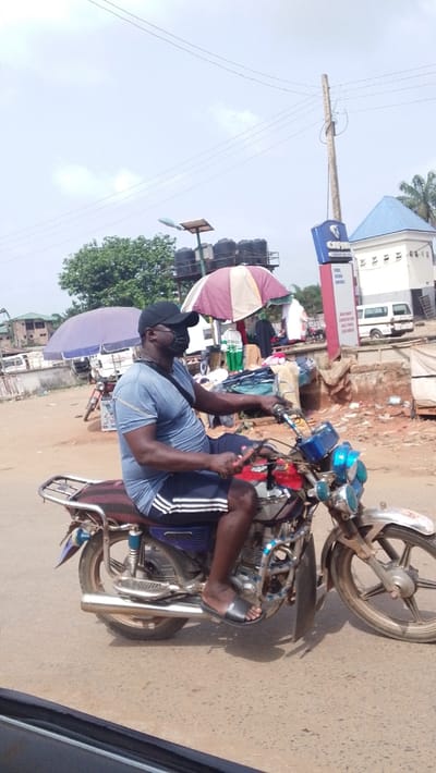 The image depicts a street scene in Igbo Ukwu, Nigeria, under an overcast sky. In the foreground, a dark-complexioned adult male is seated on a motorcycle, facing left in profile. He wears a black baseball cap, a black face mask, a light blue short-sleeved t-shirt, dark blue shorts with white side stripes, and black slide sandals. Both hands grip the motorcycle handlebars, and a dark strap extends diagonally across his chest.

The motorcycle is a standard model with red, blue, and silver components, displaying visible wear and tear, including a chrome luggage rack on the rear fender and multiple front light fixtures, some with blue tints. The rider's feet are positioned on the footrests. The road surface beneath the motorcycle is a light-colored, possibly paved or hard-packed dirt, showing some debris.

The background reveals a bustling commercial environment. To the left, a purple market umbrella is visible, alongside trees and distant multi-story buildings. Directly behind the motorcyclist, a red and white striped market umbrella shades a stall displaying various goods, possibly clothing. Above and behind this stall, several black water storage tanks are elevated on a metal frame, adjacent to a solar panel-equipped street light. Further right, a tall wooden utility pole with power lines stands next to a blue and white sign, partially readable as "OPT..." and listing "FUEL KERO DIESEL," indicating a petrol station. Behind the sign is a white building with a distinctive blue pyramidal roof, and a white van is parked in the distance.