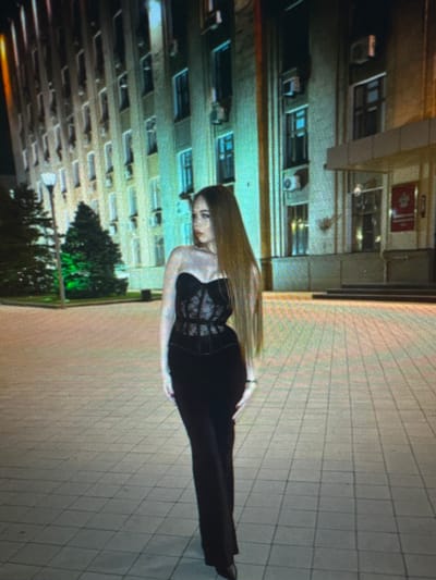 The media file depicts a woman standing on a tiled surface, possibly a sidewalk or plaza, in front of a building. The woman is wearing a black corset-like top and black pants, with her long blonde hair falling down her back. She is positioned in a way that suggests she is either posing for a photo or has just been photographed. The building behind her appears to be an apartment complex or office building with multiple windows. The time of day seems to be evening or night, as evidenced by the artificial lighting present in the scene. There are no clear indications of weather conditions. The setting could be urban, with the tiled ground and multi-story building. The image is slightly pixelated, making it difficult to discern finer details. No visible text is present in the image.