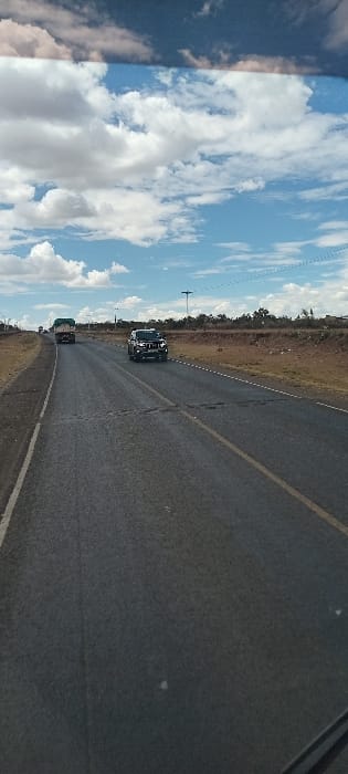 A road stretches into the distance, marked with a double yellow line. Two vehicles are visible, one following closely behind the other. The sky overhead is filled with fluffy white clouds, contrasting with the dark asphalt of the road. Vegetation lines the sides of the road, creating a border between the pavement and the open sky. A utility pole stands tall in the distance.