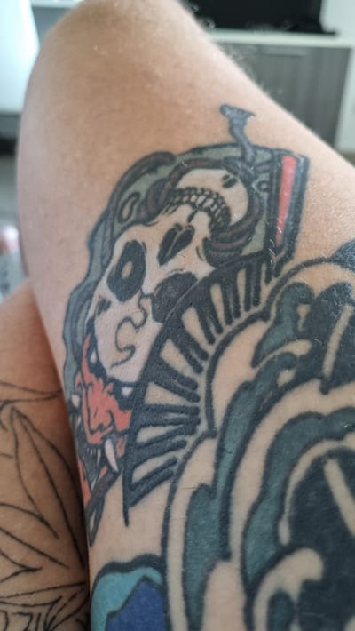 The image shows a close-up of a person's leg with a tattoo. The tattoo depicts a skull with red accents, fangs, and a fan-like design. The background is blurred and shows a hint of furniture and possibly a window, suggesting an indoor setting. There is no information available about the location being Olbia, Italy.
