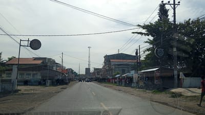 The image depicts a street scene in Palohlada, Indonesia, under a cloudy sky. A two-lane asphalt road runs through the center of the frame, flanked by buildings and various businesses. On the left side of the road, there are several low-rise structures with corrugated metal roofs, some appearing to be shops or residences. Signs and wires are visible, indicating commercial activity. A circular sign with a logo, possibly for "Vell", is mounted on a pole.
On the right side, the buildings are taller and more modern, with some featuring multiple stories. A tall tree with dense foliage stands near a utility pole, which is adorned with multiple signs, including one circular sign that reads "ESTE COFFE & DRINK". Several smaller kiosks and stalls are situated along the roadside, one displaying a "DIMSUM LIESA" advertisement.
Several vehicles, including motorcycles and cars, are present on the road, suggesting ongoing traffic. A few individuals are visible, including one person in a red shirt walking on the right side of the road and others in the distance near the shops. Telecommunication towers are visible in the background. The overall impression is that of a bustling, semi-urban environment typical of a Indonesian town.