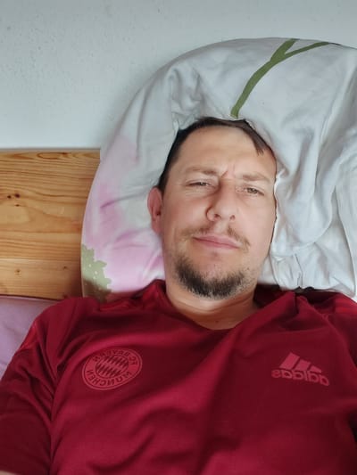 An adult male is pictured lying in a bed, looking directly at the camera. He has dark hair, a beard, and a neutral expression with a slight furrow in his brow. He is wearing a dark red short-sleeved shirt featuring the mirrored FC Bayern München crest on the left chest and a mirrored Adidas logo on the right chest, indicative of a front-facing camera shot. His head rests on a pillow with faint pink and green patterns, partially covered by a white duvet displaying a prominent green linear design. Behind him is a light wooden headboard and an off-white, textured wall. The setting is an indoor bedroom. No visual information within the image provides specific cues to confirm a location in the city of Straubing, Germany.